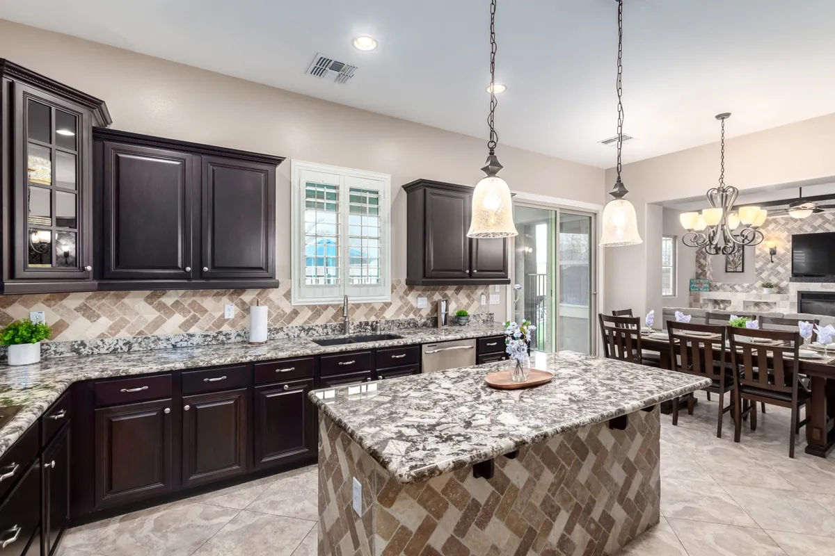 A sophisticated view of the kitchen and dining area, featuring a large granite island with dark wood cabinets, elegant light fixtures, and a dining table set beneath a chandelier. The flow between the kitchen and dining spaces evokes a sense of openness and encourages interaction.