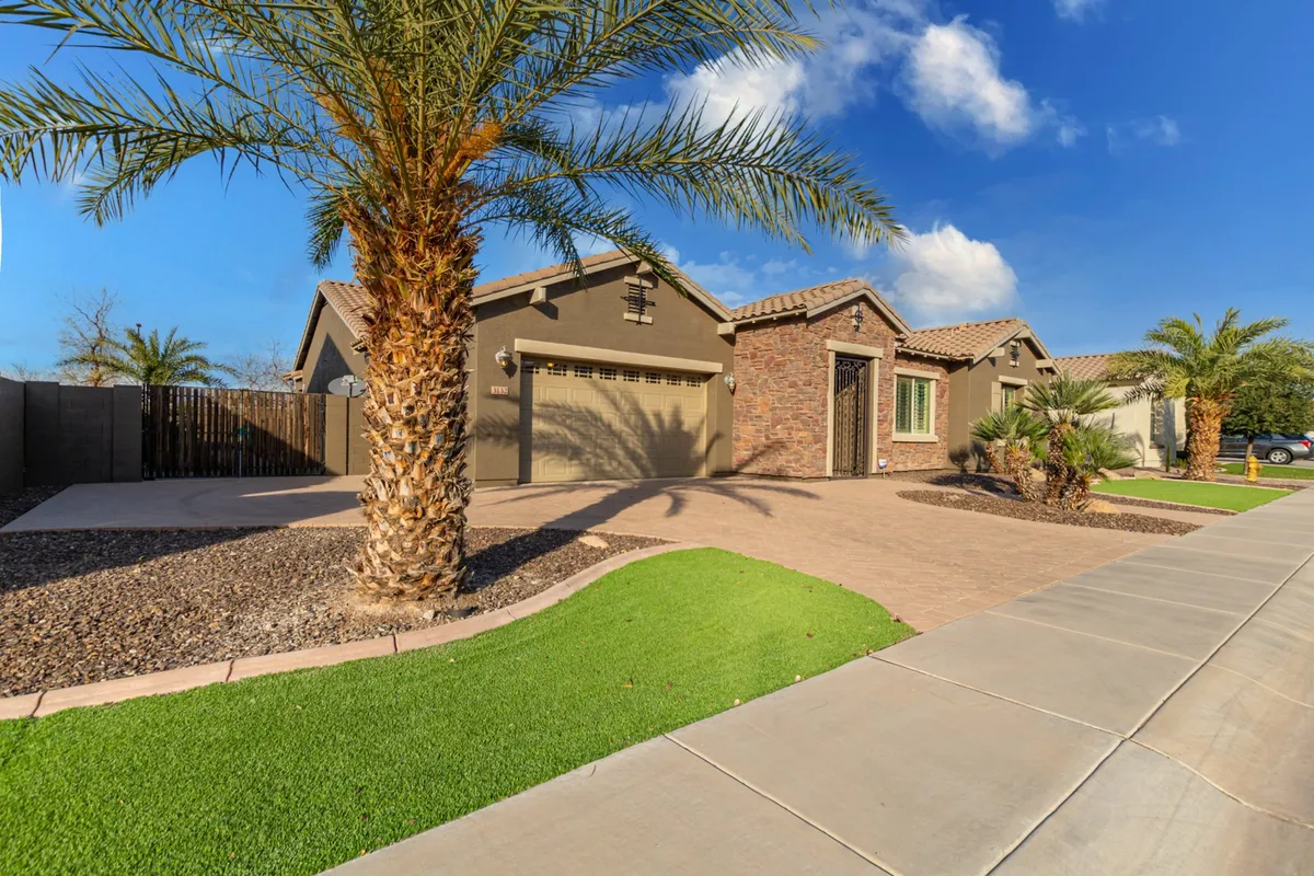 A front view of the house showcasing well-manicured lawns, tall palm trees, and a brick facade, with a paved driveway. The scene is welcoming and open, ideal for envisioning outdoor gatherings, sunbathing, or simply enjoying the Arizona weather with loved ones.
