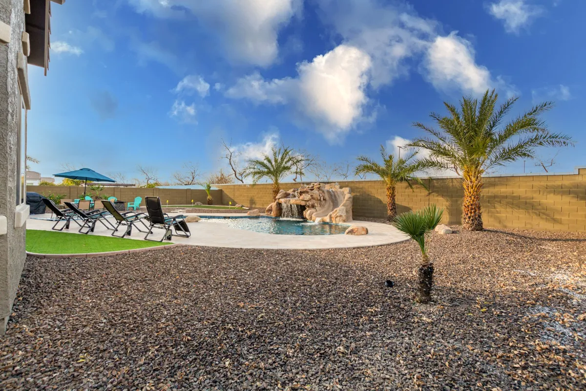 A beautifully landscaped backyard featuring a sparkling swimming pool with a rock waterfall, surrounded by palm trees and lounge chairs. An outdoor umbrella provides shade, creating an inviting space for relaxing or socializing by the water.