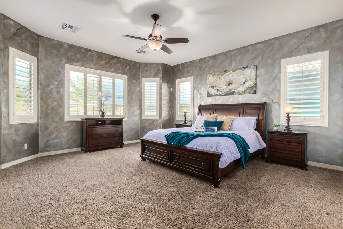A serene bedroom featuring a comfortable bed adorned with decorative pillows, flanked by nightstands with lamps. Artwork decorates the wall and shuttered windows let in soft, natural light, creating a tranquil, restful atmosphere.