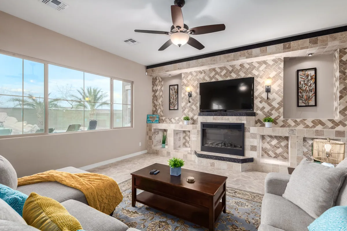 A spacious great room featuring a cozy seating arrangement with sofas and a coffee table, all oriented towards a fireplace and large TV. The area is flooded with natural light from large windows, and there’s a seamless open flow that visually connects this living area with the adjoining spaces.