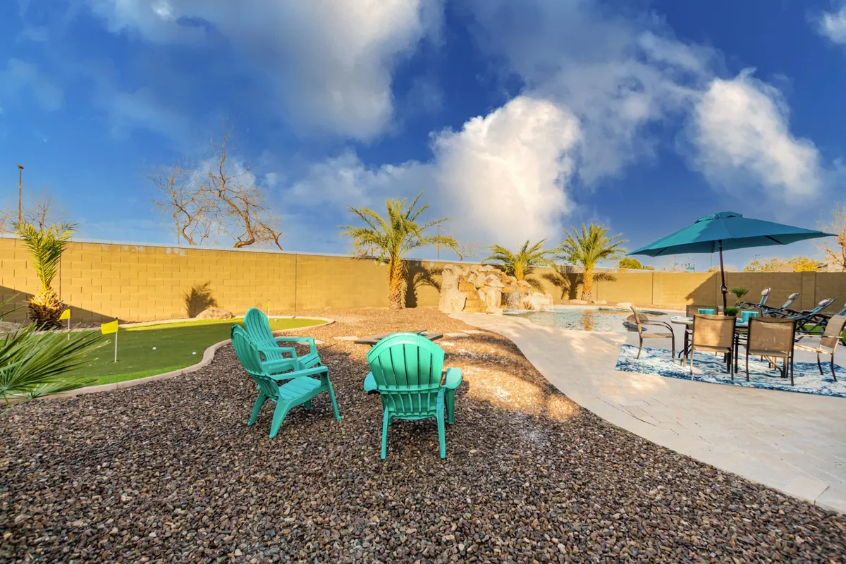 A tranquil backyard scene featuring turquoise chairs overlooking a pool area, palm trees, and a rock feature. The space is designed for both relaxation and socializing, with clear views of the putting green and space for outdoor games.