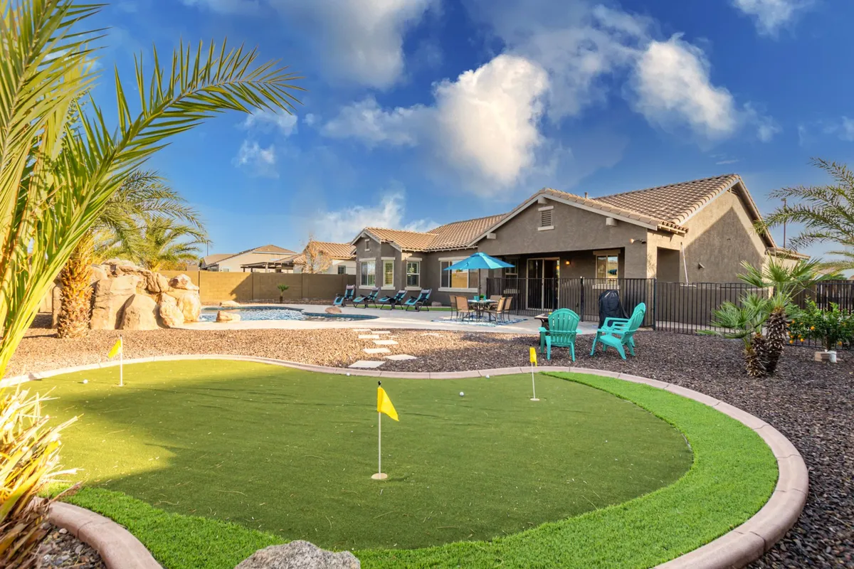 A lively backyard scene featuring a well-maintained putting green adjacent to the swimming pool. The space is surrounded by lush landscaping, palm trees, and offers inviting outdoor seating areas. The overall mood is cheerful and family-friendly, suggesting opportunities for both play and relaxation.