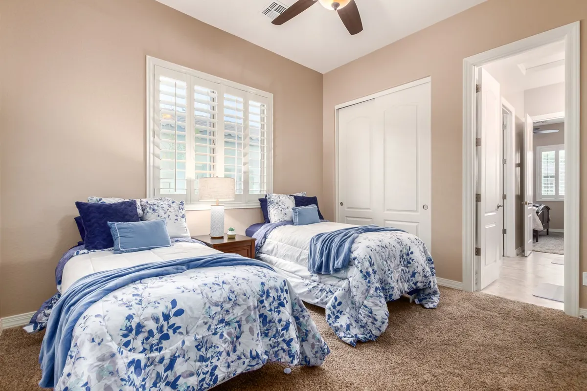 A bright and cozy bedroom with two twin beds featuring floral bedding, a central nightstand with a lamp, and a large window with plantation shutters letting in daylight. The room feels flexible, perfect for families or groups needing adaptable sleeping arrangements.